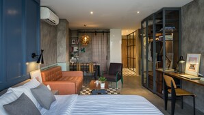 Une chambre d’hôtel moderne avec un lit, un canapé, un bureau et une chaise. On y trouve une petite table avec une plante et une lampe. La chambre a un mur bleu et une armoire en bois.