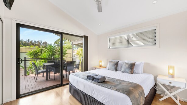 A bedroom with a large bed, a nightstand, a ceiling fan, and a sliding glass door leading to an outdoor patio with a table and chairs. - Mountain View Resort (Shoalhaven Heads)