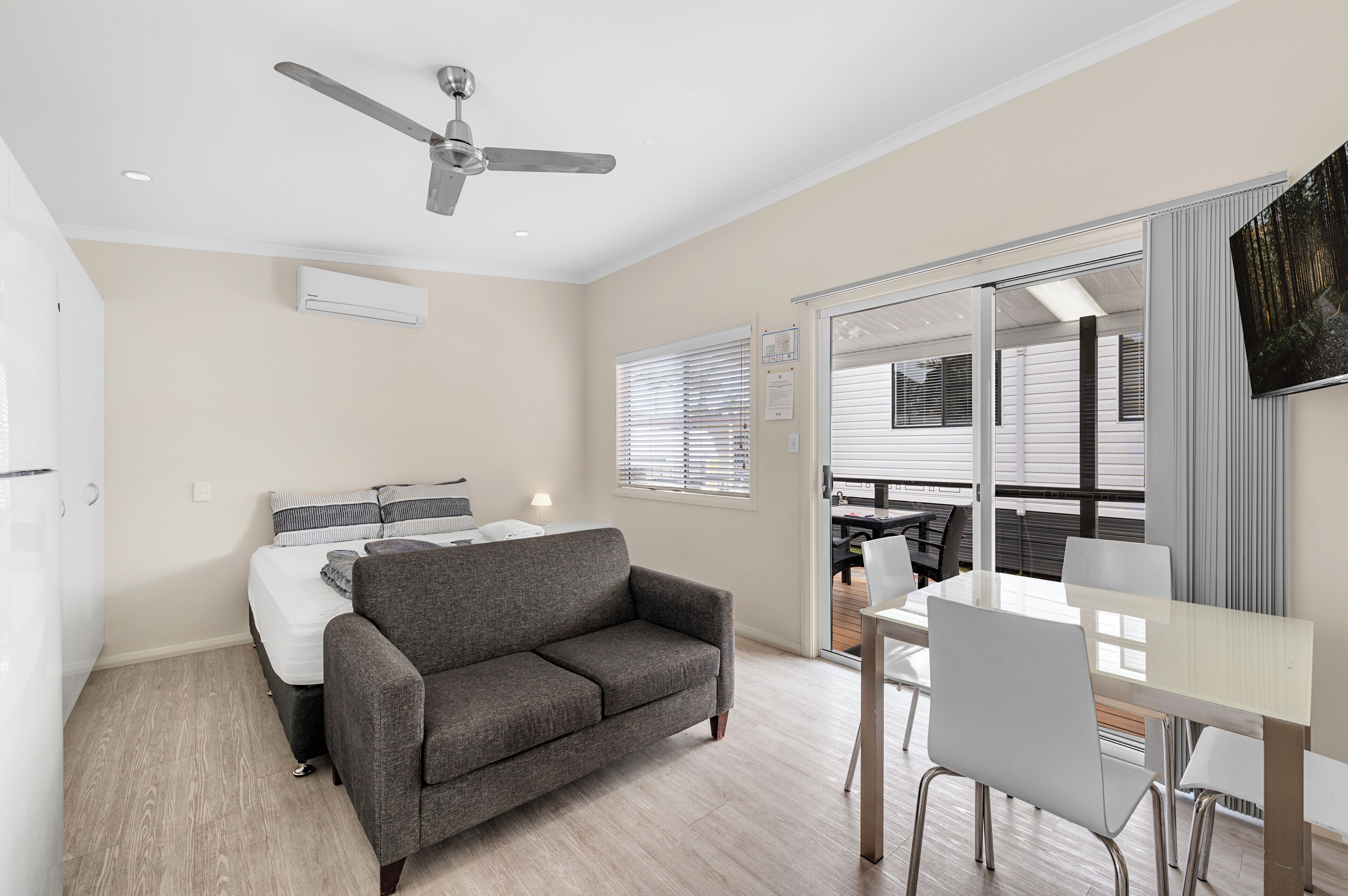 A modern bedroom with a bed, a gray sofa, a dining table with chairs, a ceiling fan, and a sliding glass door leading to a balcony.
