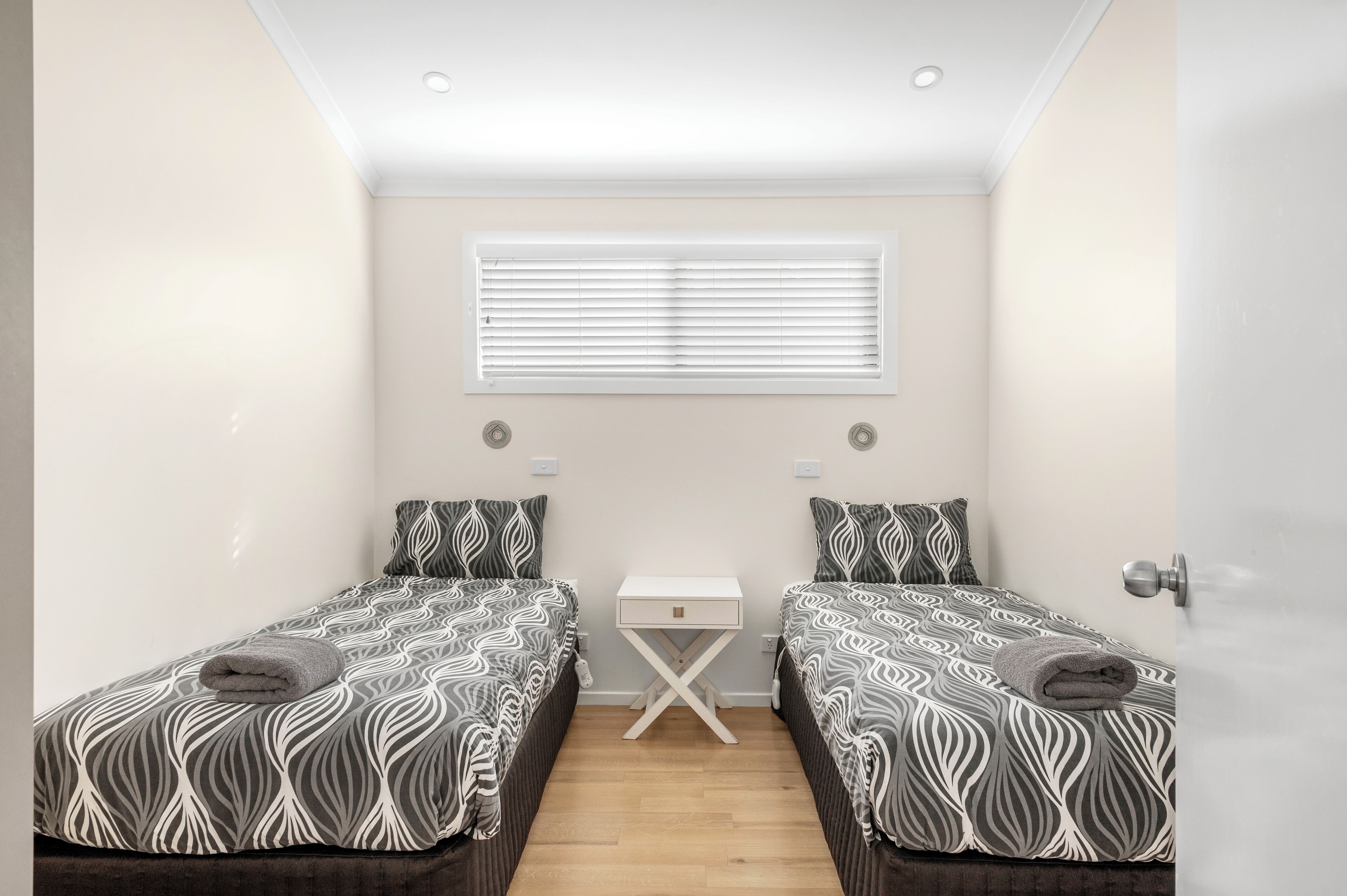A small, well-lit bedroom with two beds, a bedside table, and a window with blinds.