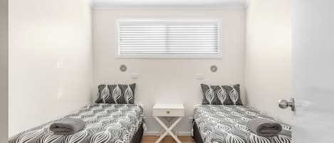 A small, well-lit bedroom with two beds, a bedside table, and a window with blinds.