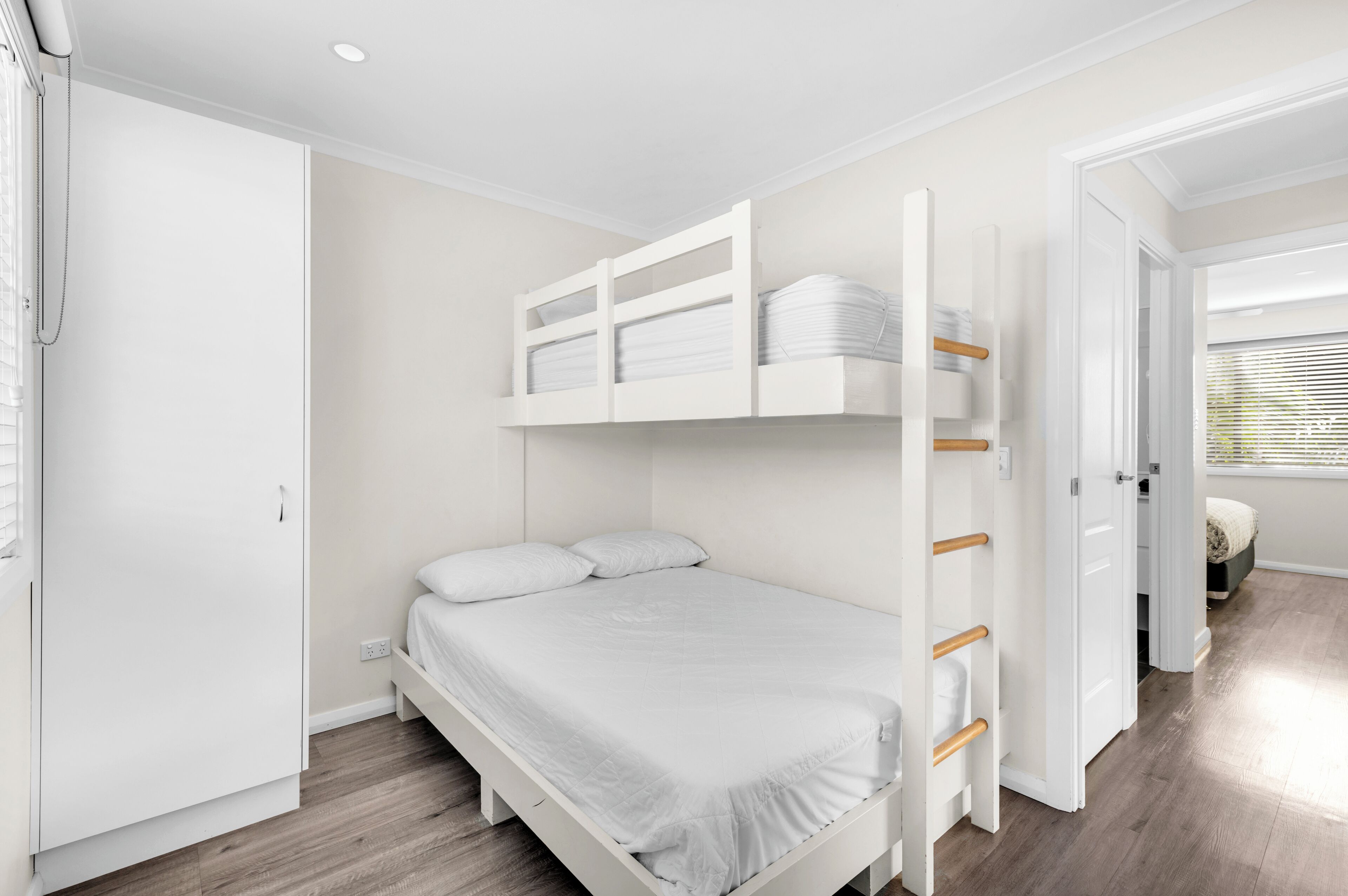 A bunk bed with a white mattress and pillows, a white wardrobe, and a wooden floor.