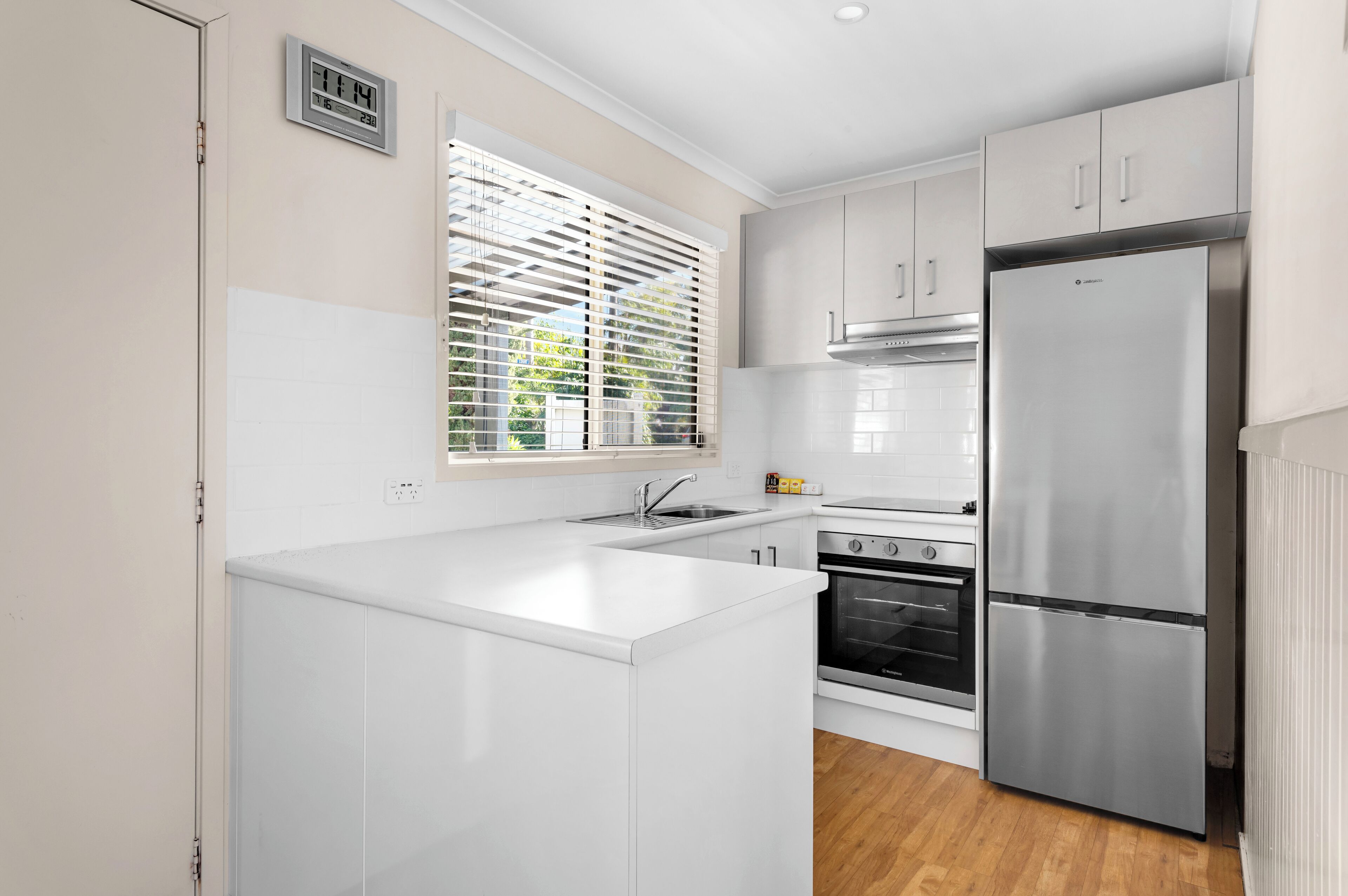 A modern kitchen with white cabinetry, a stainless steel refrigerator, a built-in oven, and a window with blinds.