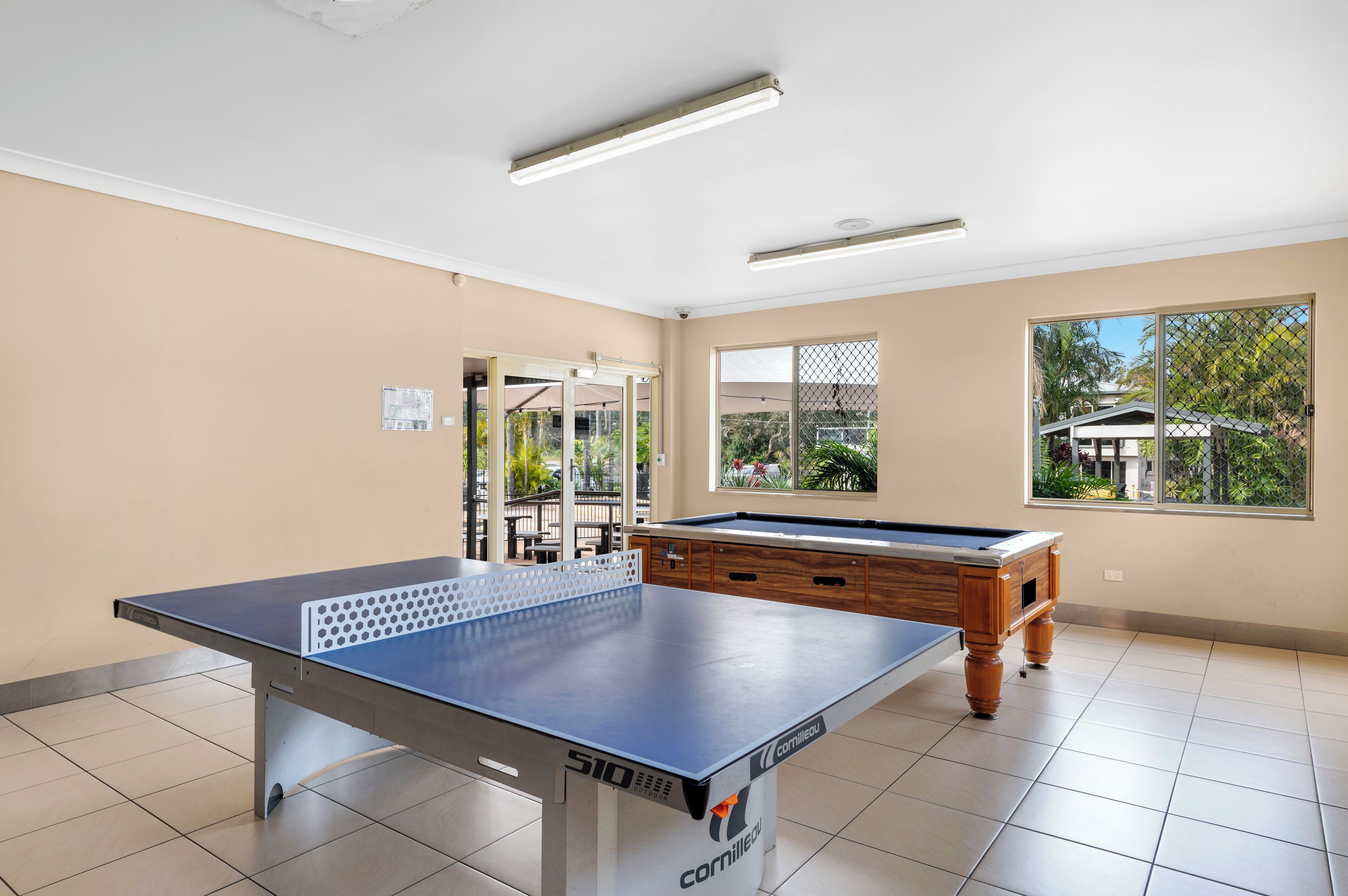A billiards table with a pool cue, a wooden pool table, and a window with a view of greenery.