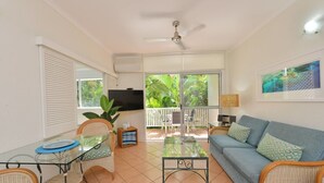 A living room with a glass table, wicker chairs, a sofa, a TV, and a balcony with a view of greenery.