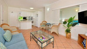 A living room with a sofa, chairs, a coffee table, a TV, and a kitchen area with cabinets and a dining table.