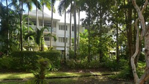 A hotel with a white exterior, multiple balconies, and lush tropical vegetation surrounding the building.