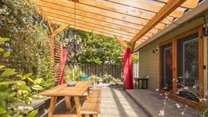 En pergola av trä med ett bord och en bänk, en trädgård med växter och en liten staty, och ett hus med en skjutdörr i glas.