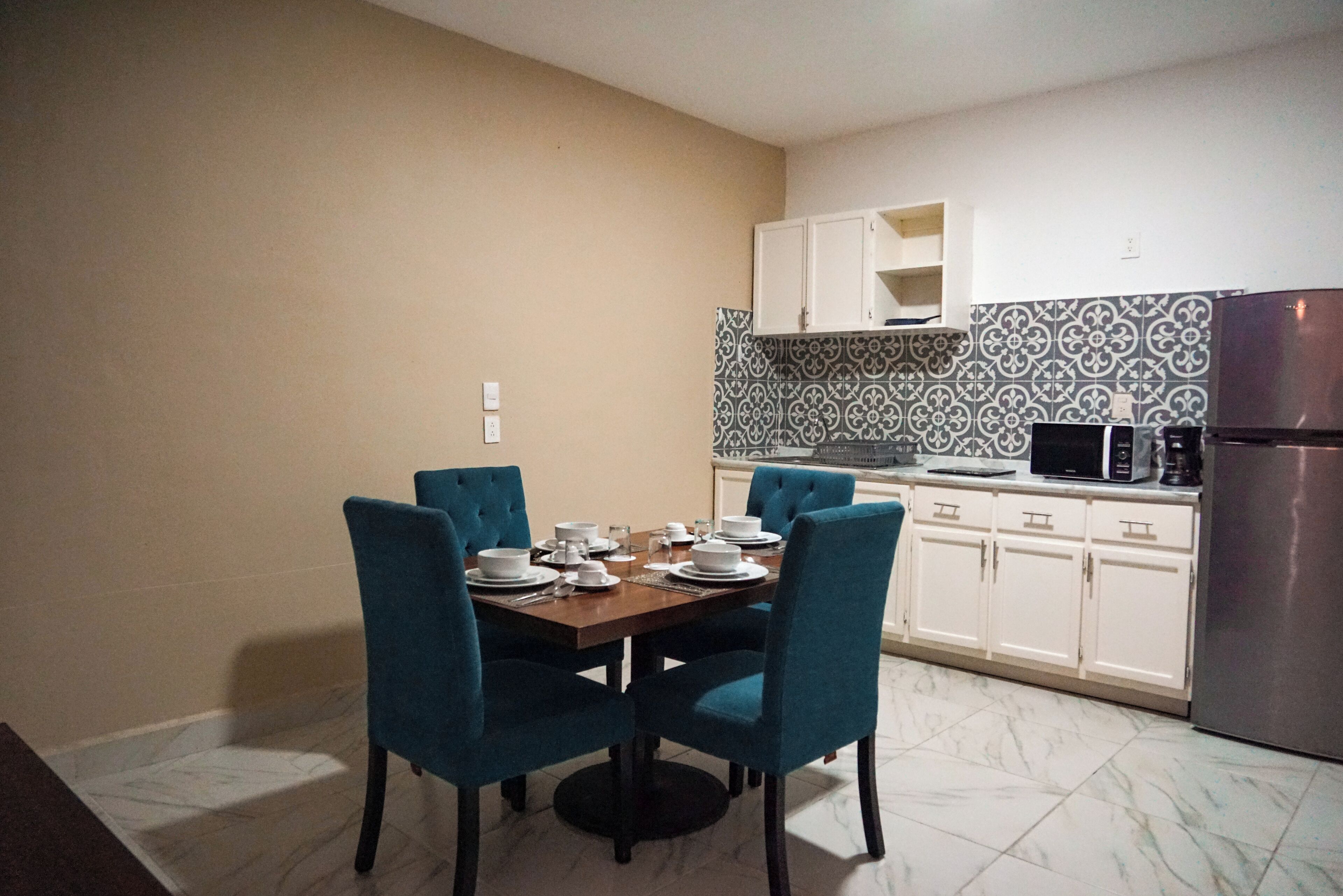 Un comedor con una mesa dispuesta para cuatro, sillas azules, armarios blancos, un microondas y un refrigerador.