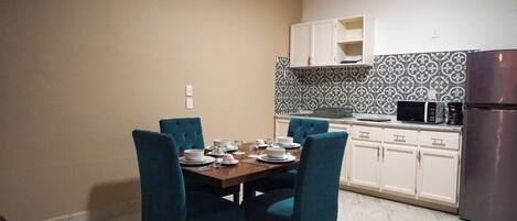 Un comedor con una mesa dispuesta para cuatro, sillas azules, armarios blancos, un microondas y un refrigerador.