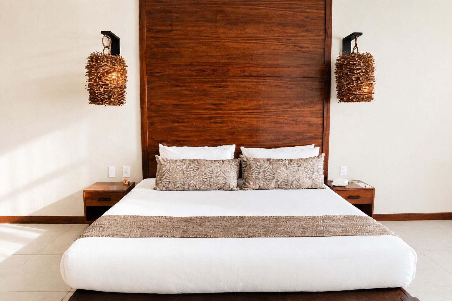 Une chambre d’hôtel avec une grande tête de lit en bois, un lit avec une literie blanche et à motifs, deux tables de chevet et des appliques murales.
