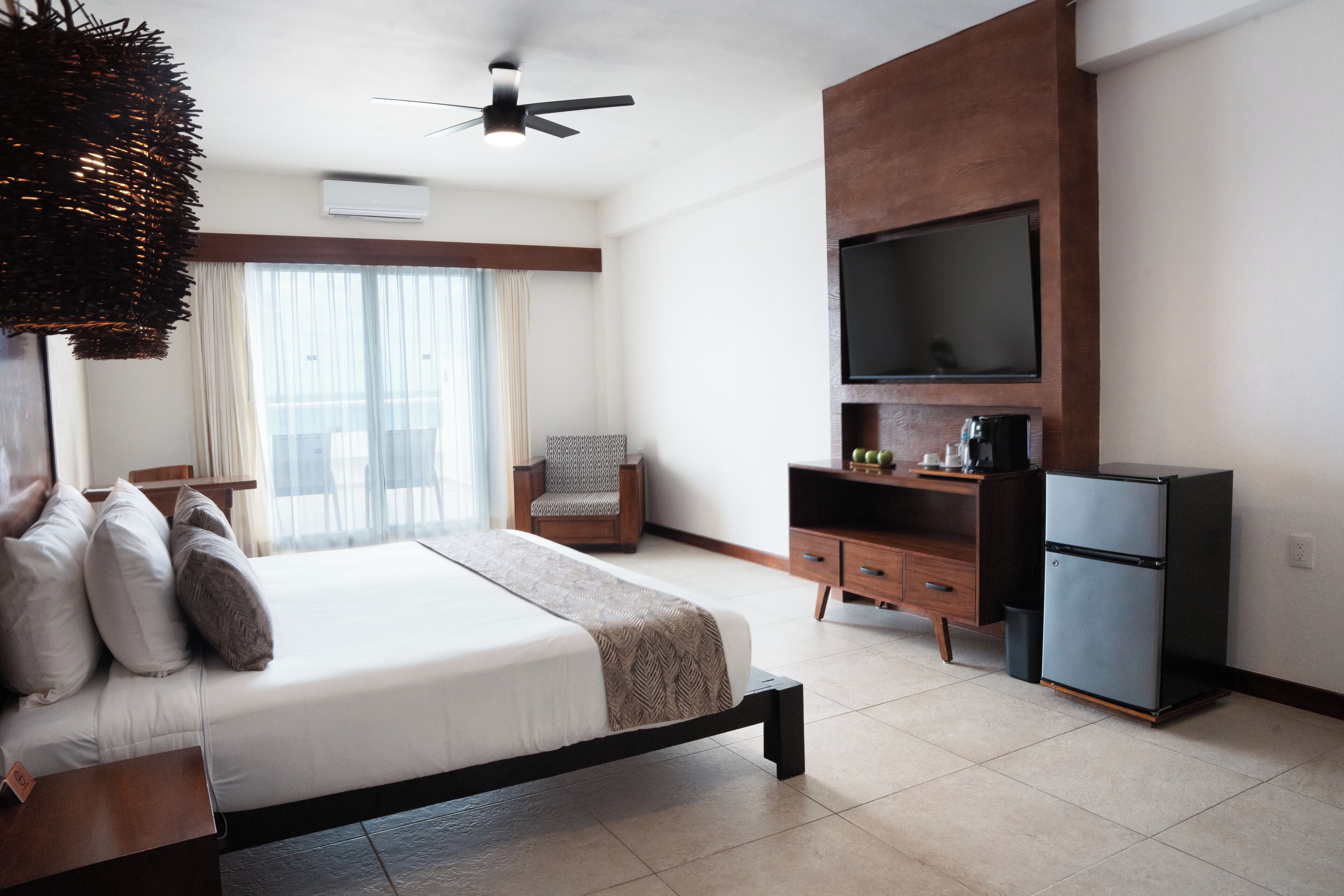 Habitación de hotel con cama, una silla, televisor, refrigerador y ventilador de techo.