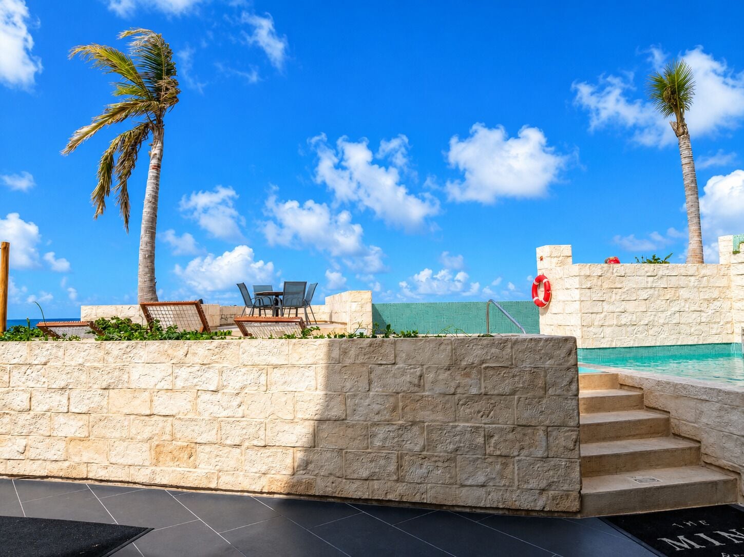 A pool area with stone walls, palm trees, and a clear blue sky.