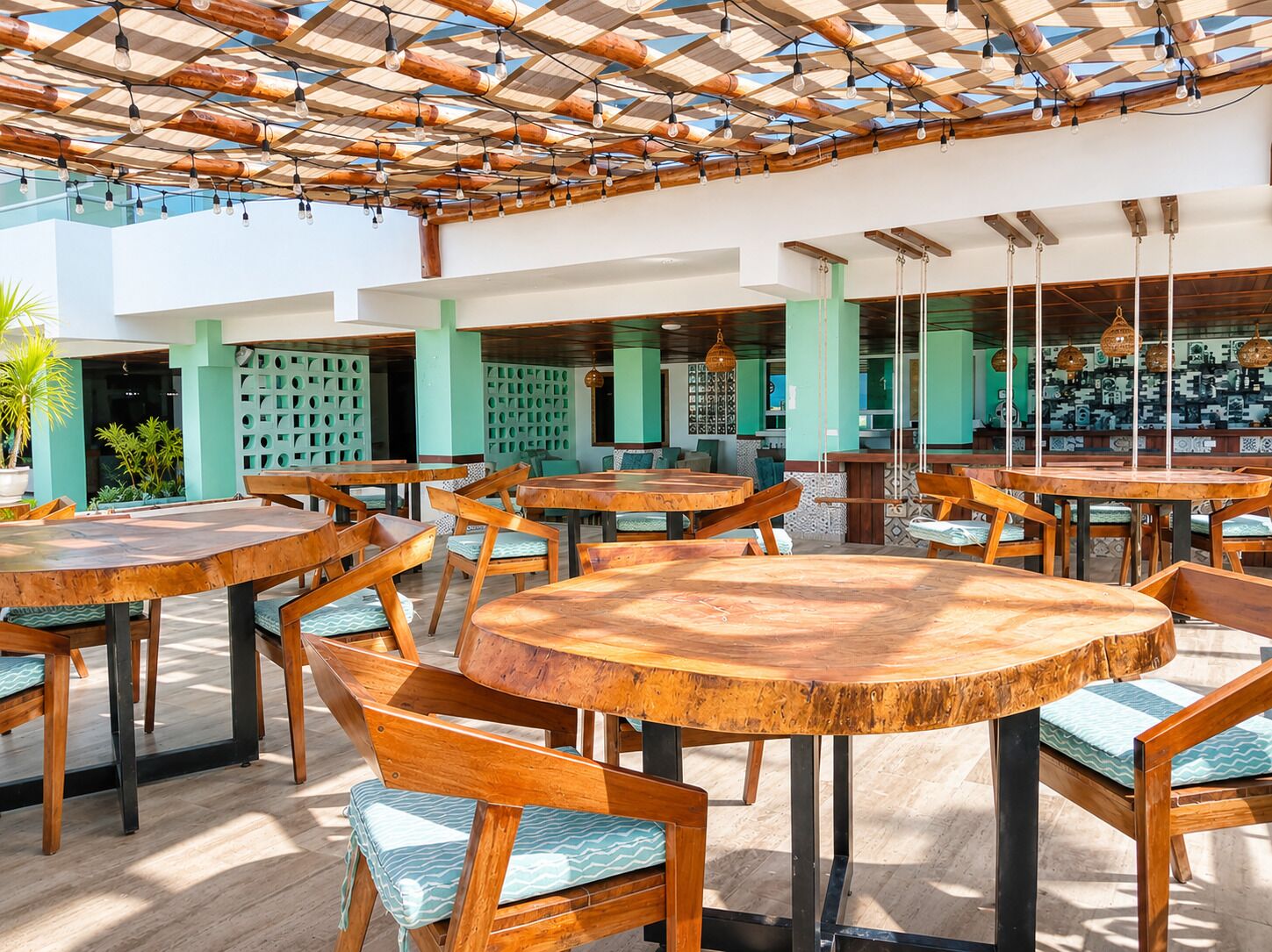 Un área de comedor al aire libre con mesas y sillas de madera, techo de madera con luces colgantes y vista al interior a través de paredes de vidrio.
