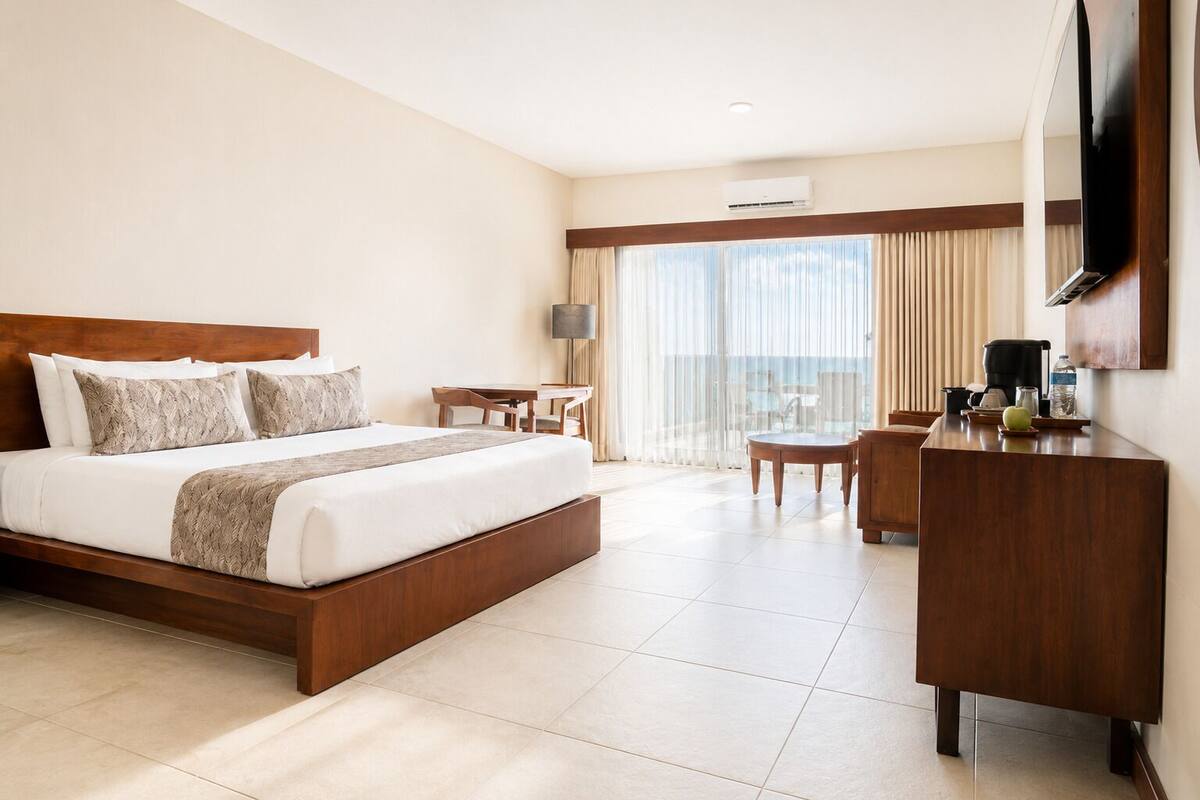 A hotel room with a large bed, a desk, a chair, a TV, and a view of the sea.