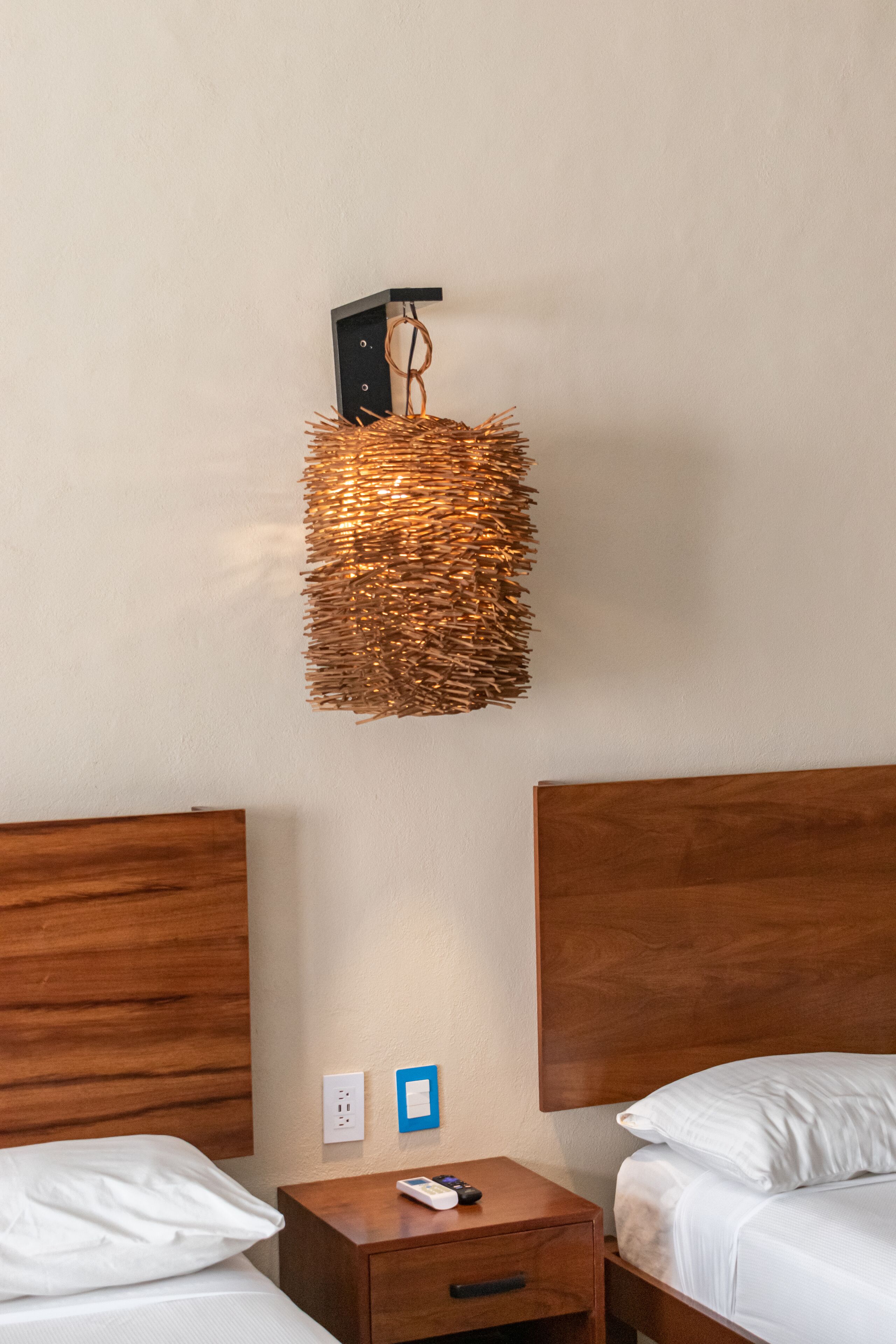 Habitación de hotel con dos camas, cabeceros de madera, una mesita de noche con control remoto y una lámpara de pared.