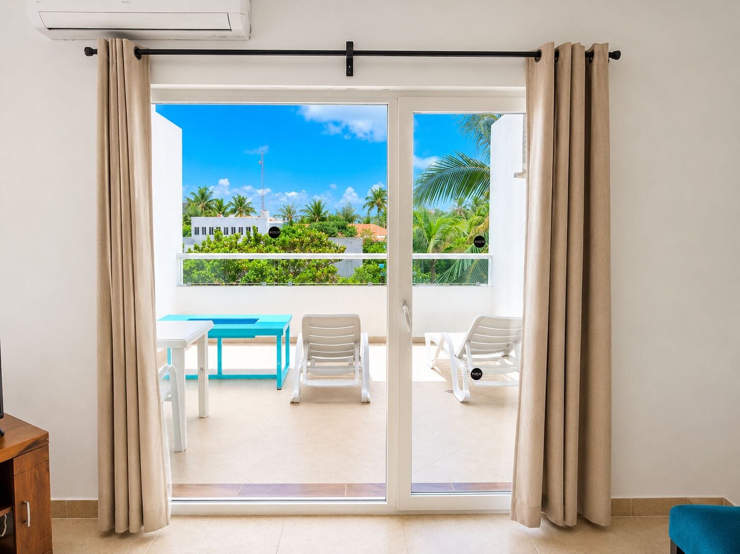 A room with a sliding glass door leading to an outdoor balcony with a table and chairs, a view of greenery and buildings, and an air conditioning unit on the wall.