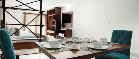 A dining area with a table set for four, a sofa, a TV mounted on the wall, and a wooden structure in the background.