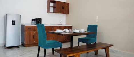 A compact kitchen with a refrigerator, a coffee maker, and a sink. There is a dining table with two chairs and a bench.
