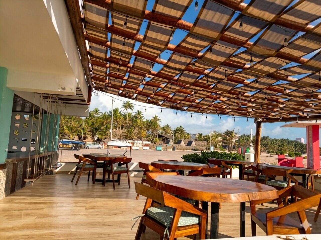 Een buitenzitgedeelte met houten tafels en stoelen, een overdekt dak en uitzicht op palmbomen en een strand.
