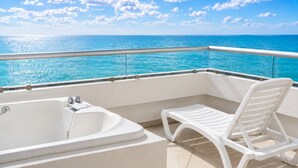 Un balcón con una bañera blanca y una silla lounge blanca, con vistas al mar turquesa bajo un cielo despejado.