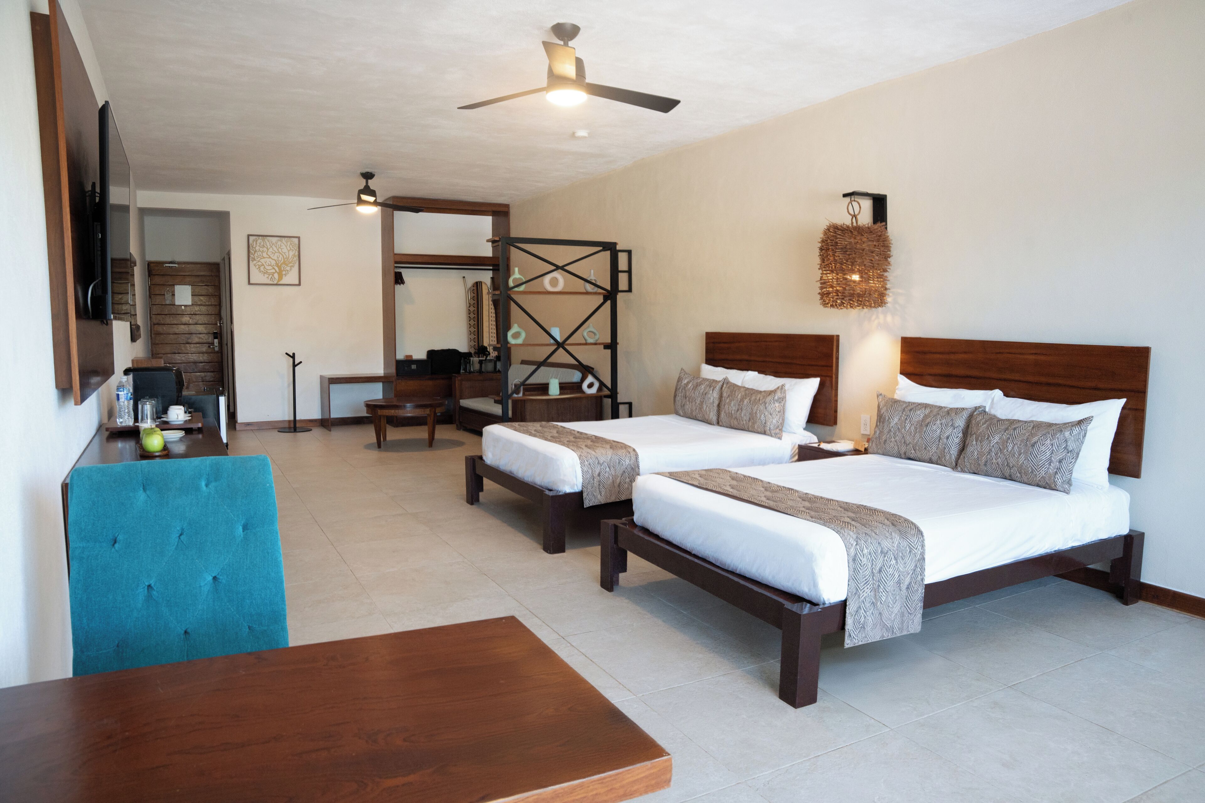 A hotel room with two beds, a wooden desk, a blue chair, a ceiling fan, and a wall-mounted TV.