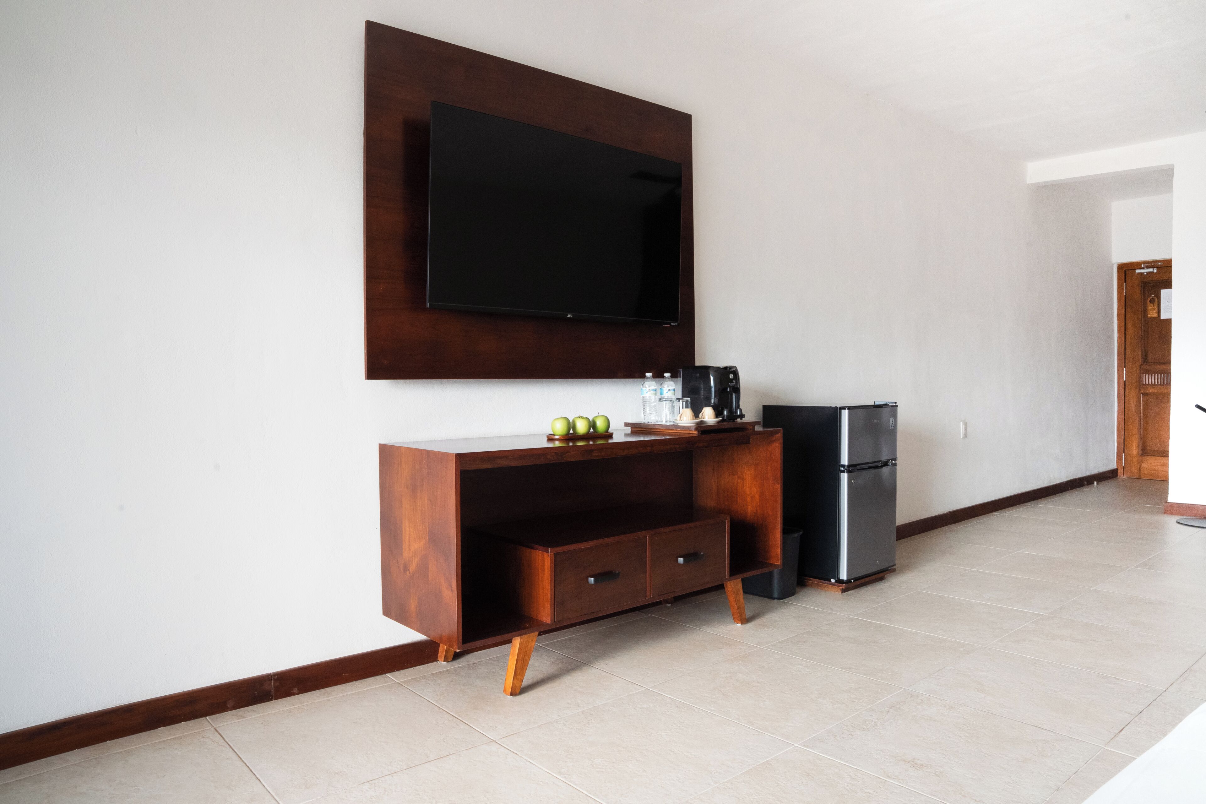 A modern living room with a flat-screen TV mounted on the wall, a wooden cabinet with drawers, a small refrigerator, and a tiled floor.