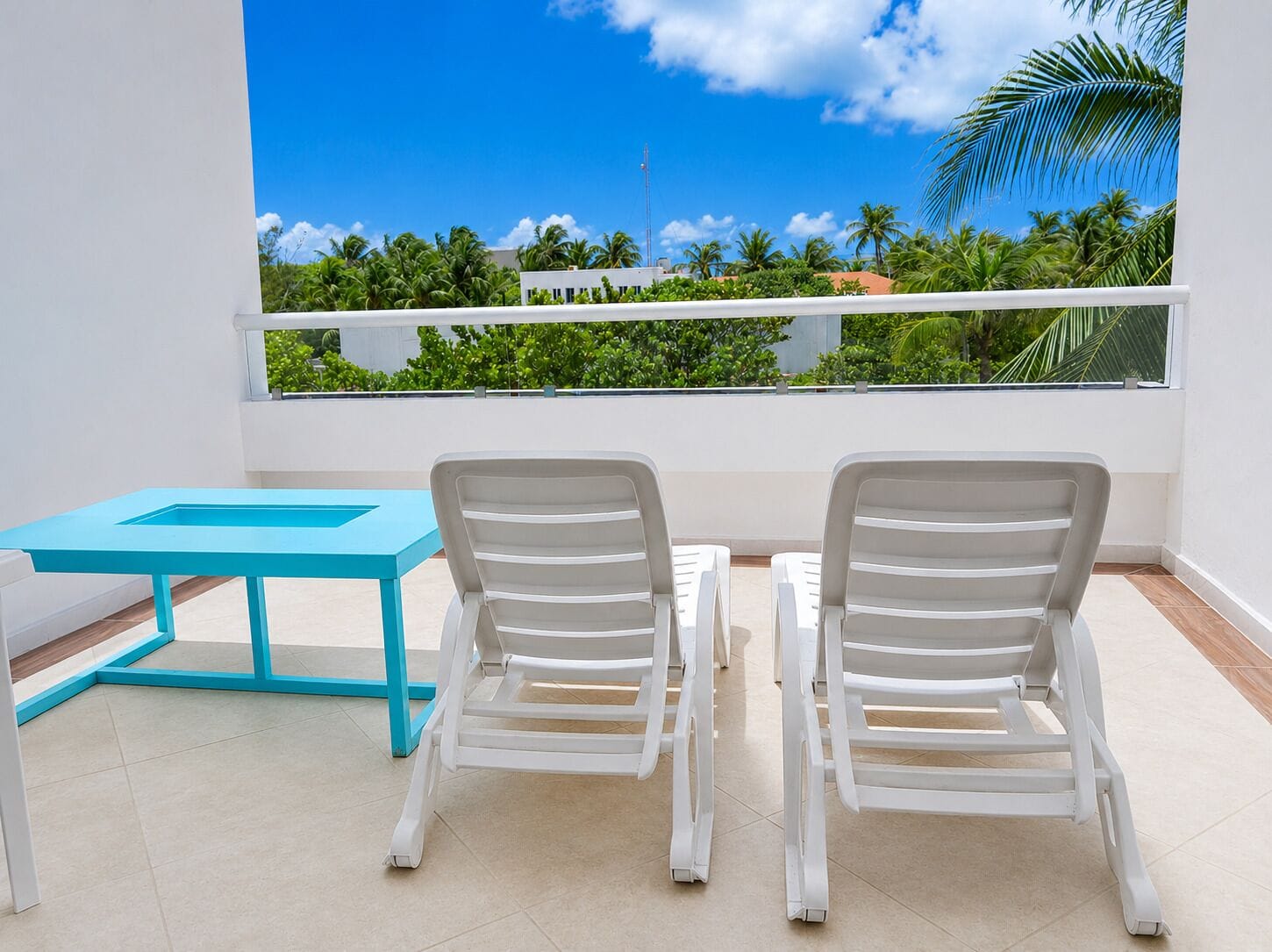 Een balkon met twee witte stoelen, een turquoise tafeltje en uitzicht op palmbomen en gebouwen.
