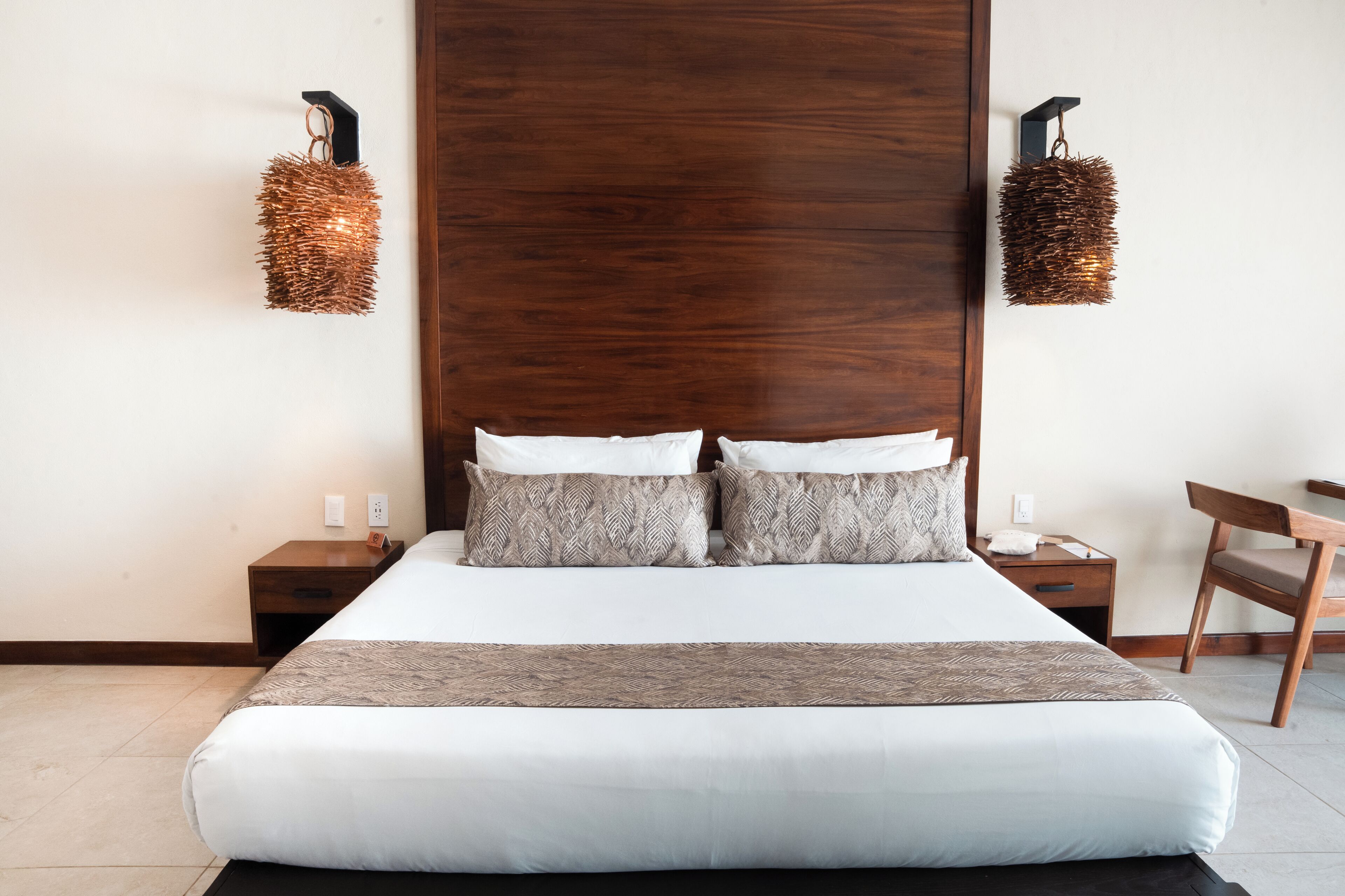 Un lit bien fait, avec une tête de lit, deux tables de chevet, une chaise et deux lampes fixées au mur.