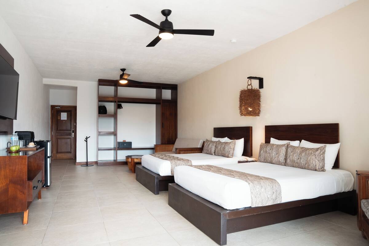 A hotel room with two beds, a desk, a chair, a ceiling fan, and a wall-mounted light fixture.