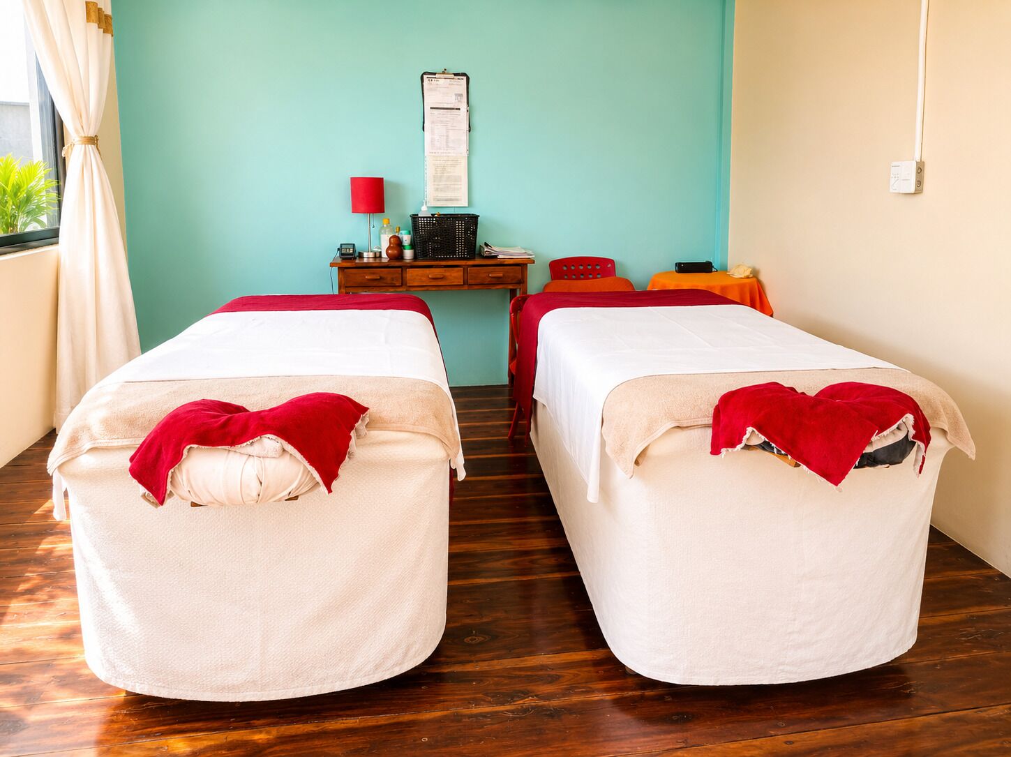 A room with two beds, a desk, a chair, a red lamp, and a wall-mounted item.