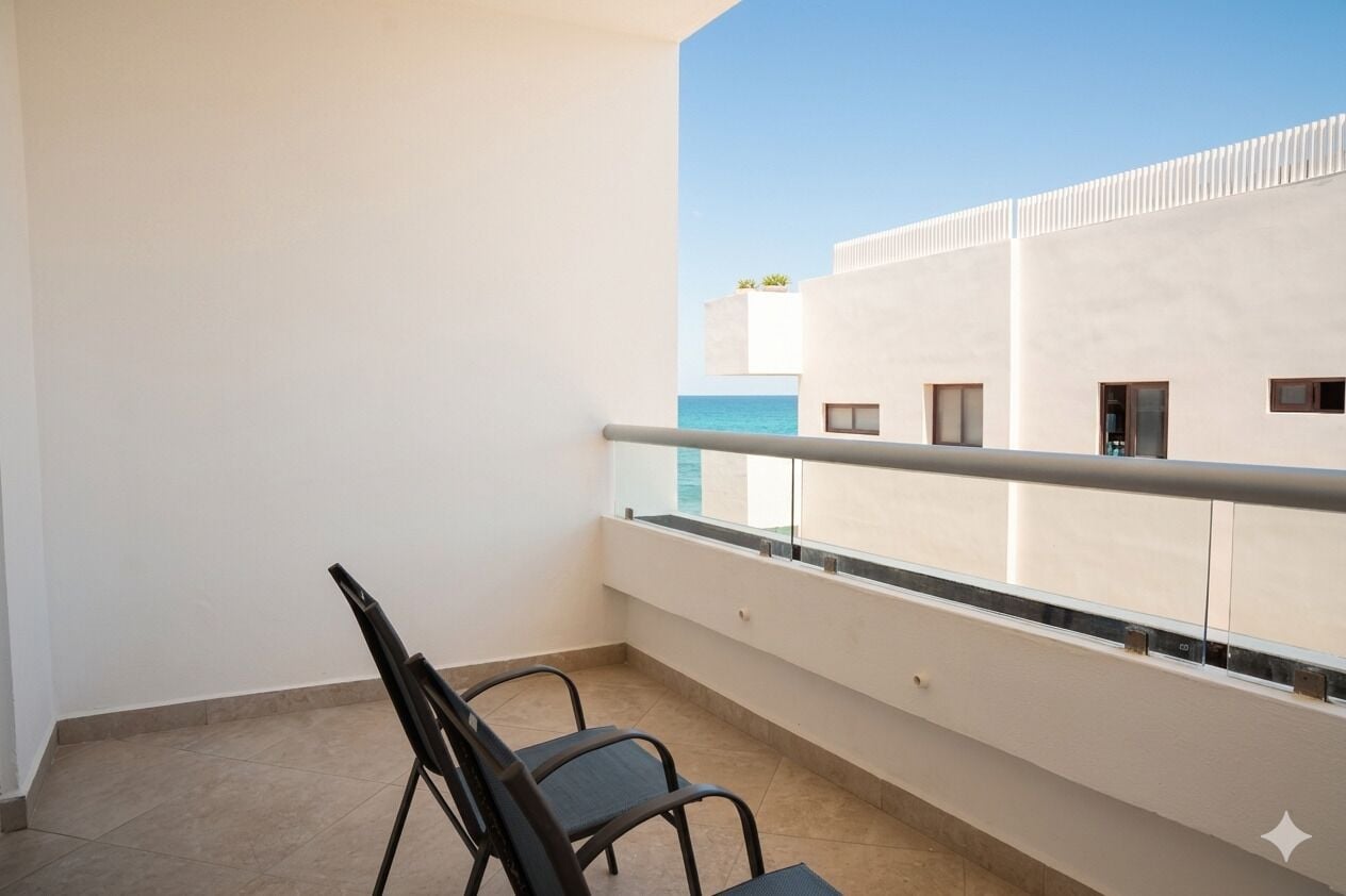 Un balcón con barandilla de metal, una silla y vistas de un edificio con techo plano y ventanas.