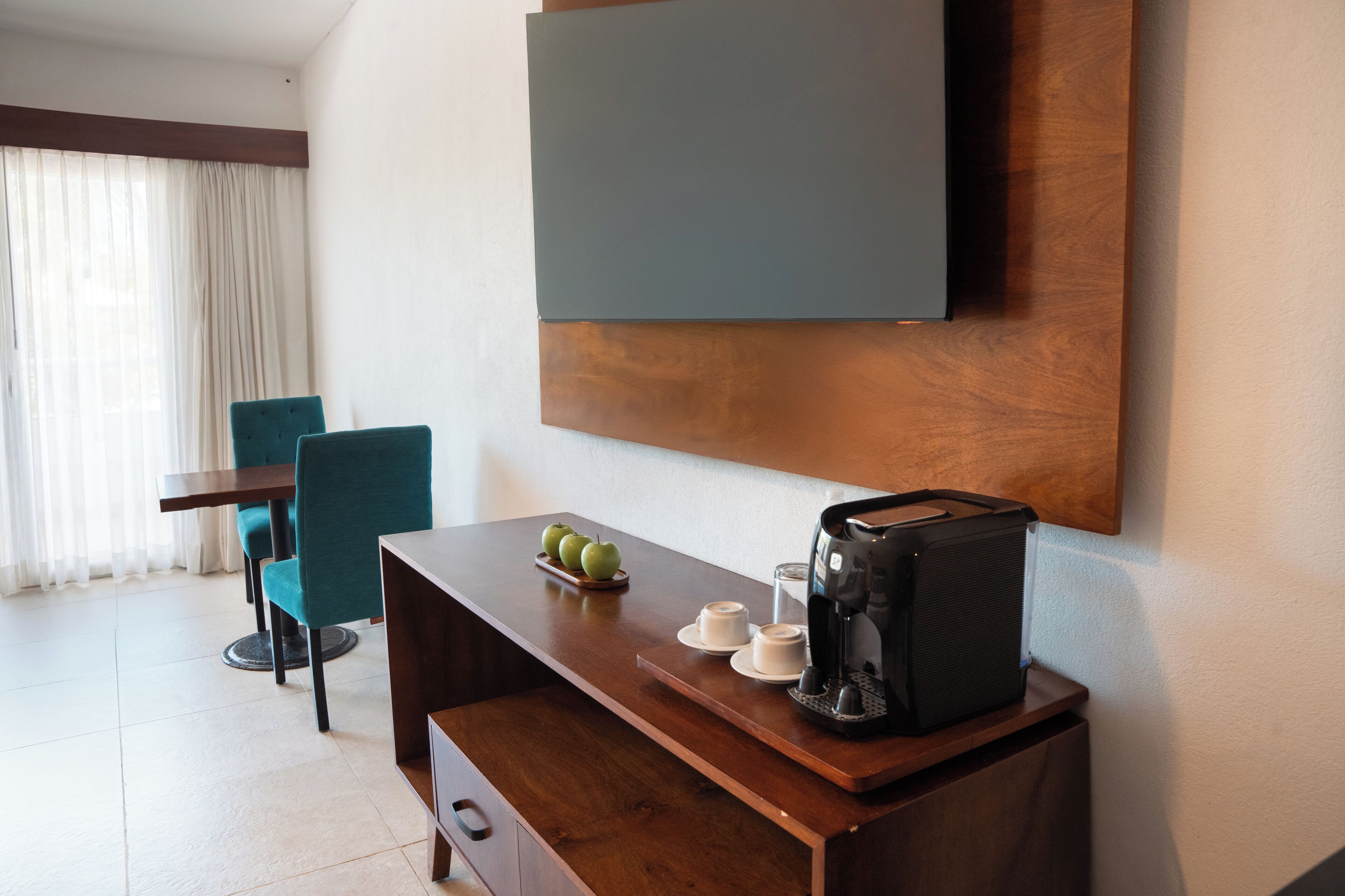 A hotel room with a wooden desk, a coffee maker, a plate of apples, and a TV mounted on the wall.