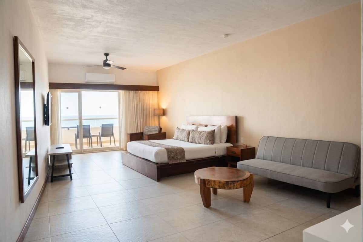 A hotel room with a bed, a sofa, a round wooden coffee table, a desk, and a chair. There is a balcony with a view.