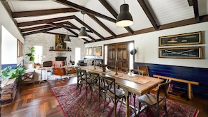 A spacious dining area with a long wooden table, multiple chairs, and a red patterned rug. The room features exposed wooden beams on the ceiling, stone walls, and framed artwork on the walls.