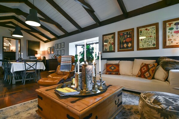 A cozy living room with a leather chair, a wooden coffee table, and a sofa. There are framed botanical prints on the wall and a dining area with a table and chairs.