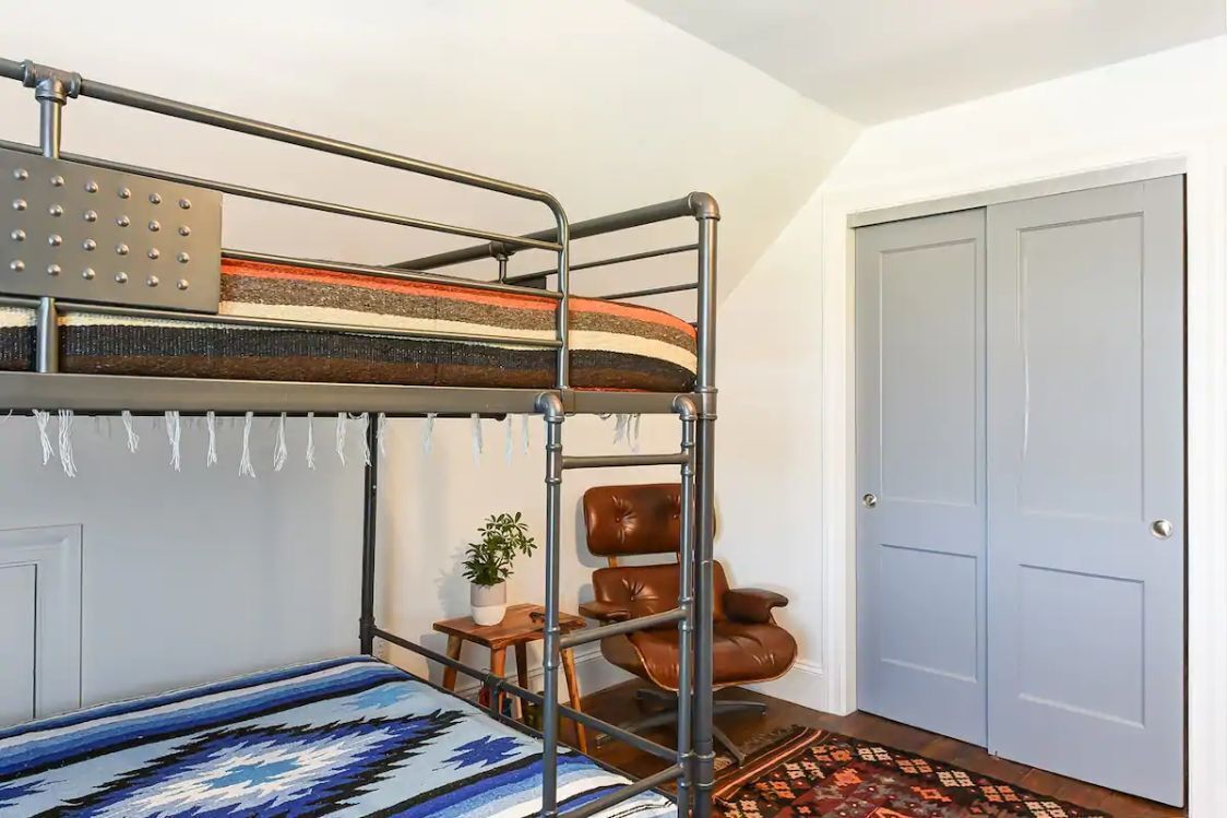 Een stapelbed met een metalen frame, een bruine lederen fauteuil, een bijzettafeltje met een plant en een kamerbreed tapijt met een patroon.