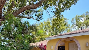 Un edificio amarillo con tejado de baldosas, una entrada arqueada y un jardín con diversas plantas y un árbol.