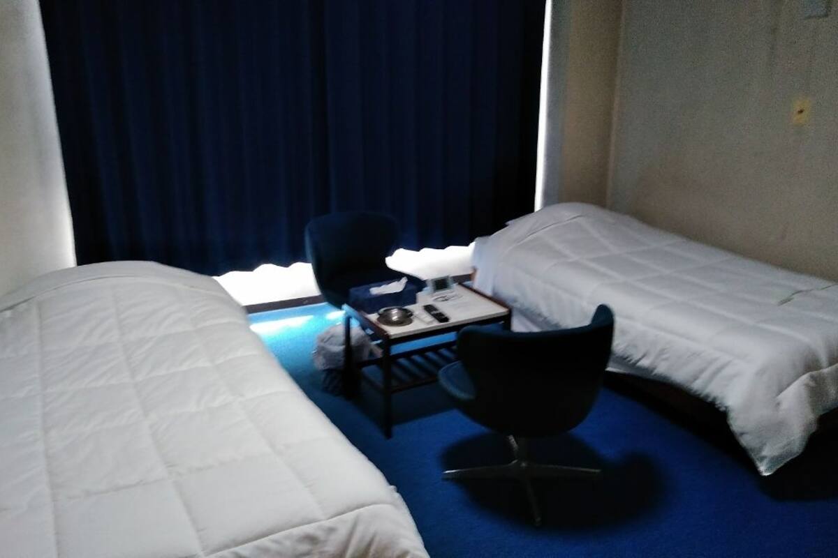 A small hotel room with two single beds, a desk with a chair, and a window with curtains.
