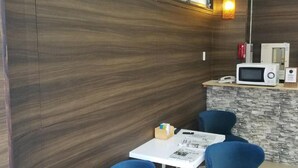 A small, modern room with a wooden wall, a white table, two blue chairs, a small wooden shelf with papers, and a microwave on a counter. - Hotel Takanoya (Hadano)