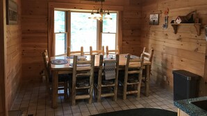 Interior de una cabaña de madera con una mesa de comedor y sillas, una araña y una ventana con vista a los árboles.