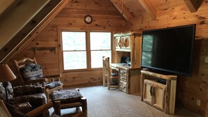 Un interior de cabaña acogedor con un televisor grande, muebles de madera y un ventanal que deja entrar luz natural.