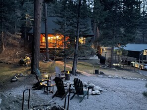 A lodge with a fire pit area, outdoor seating, and a large illuminated building in the background. - Towering Pines Cabin (Beulah Valley)
