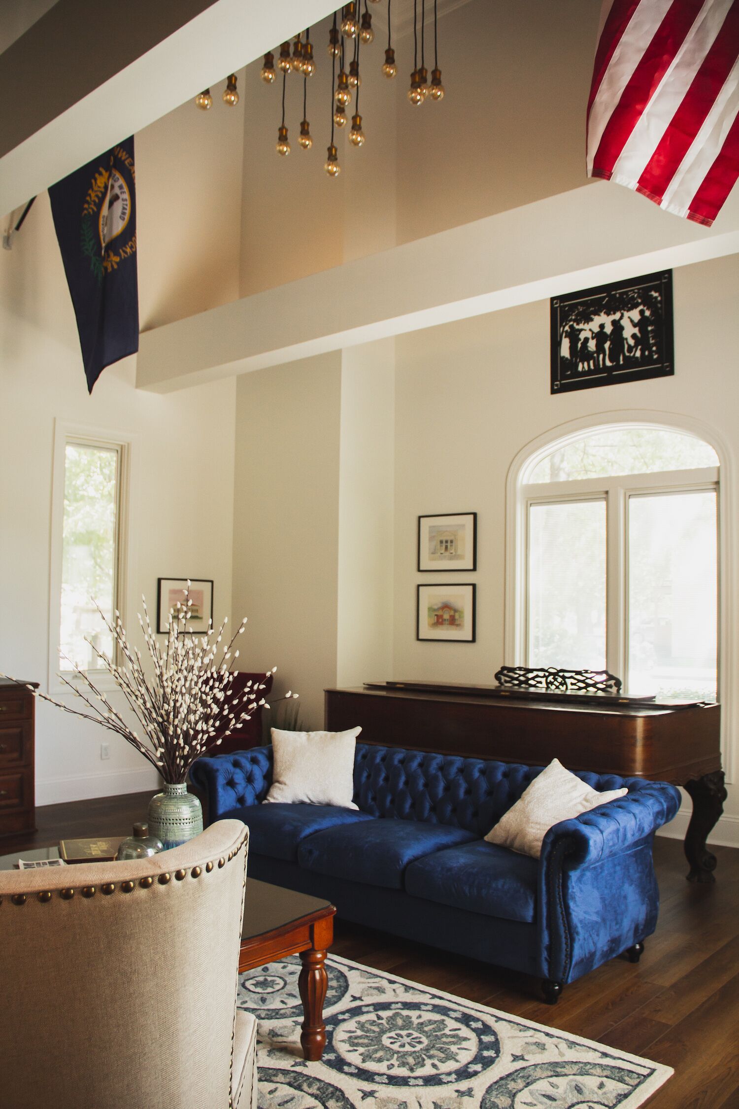 Un salon avec un canapé bleu, une table basse en bois et un vase décoré de branches. Un drapeau est accroché au mur et une fenêtre offre une vue sur l’extérieur.