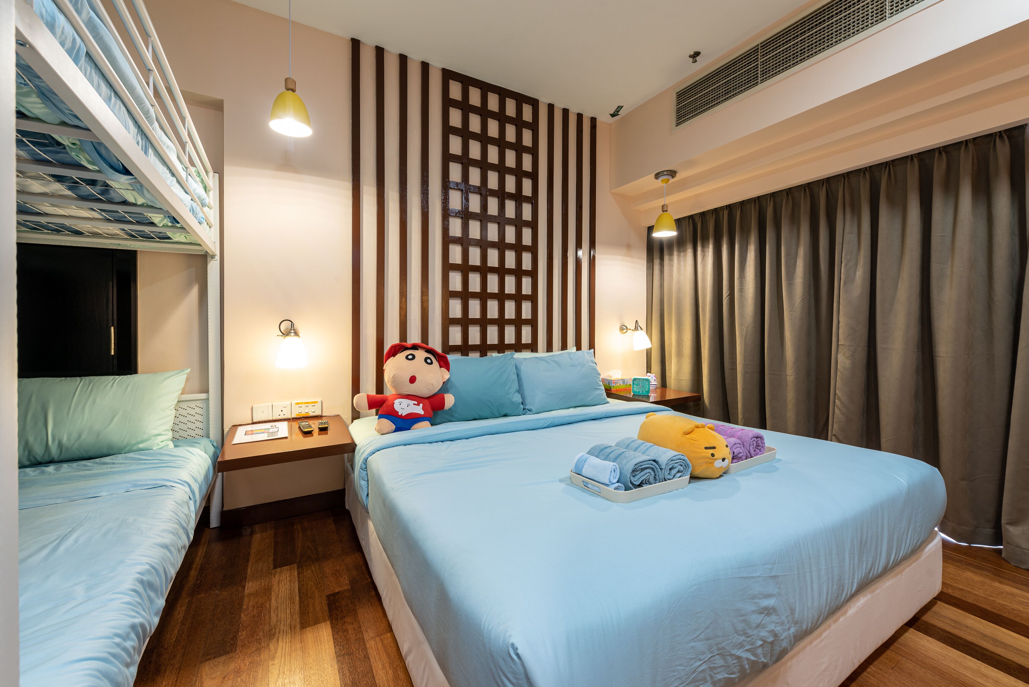 A hotel room with a bunk bed, a large bed with a blue bedspread, a wooden floor, a decorative wall panel, and a window with curtains.