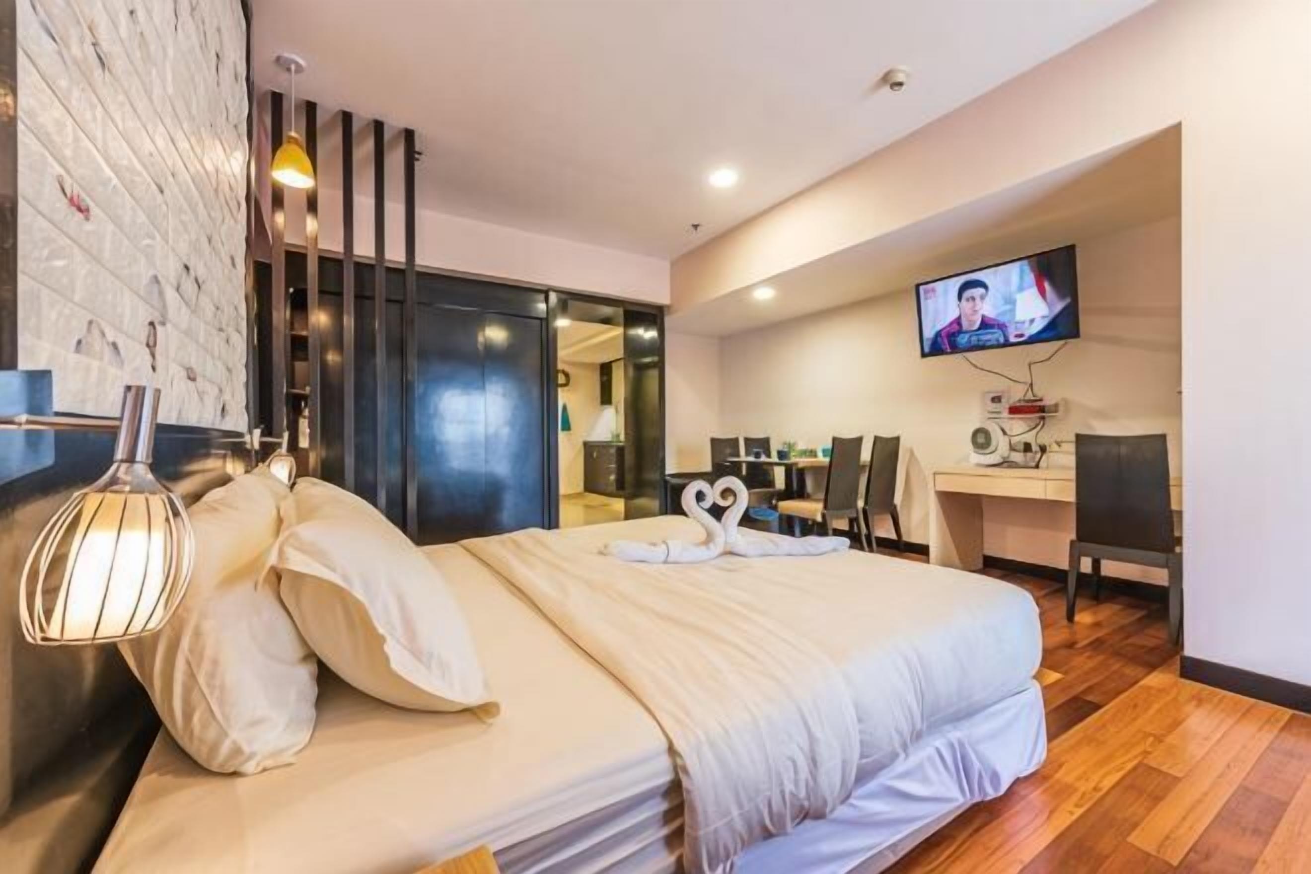 A modern hotel room with a large bed, a TV, a desk with a chair, and a wall-mounted TV.