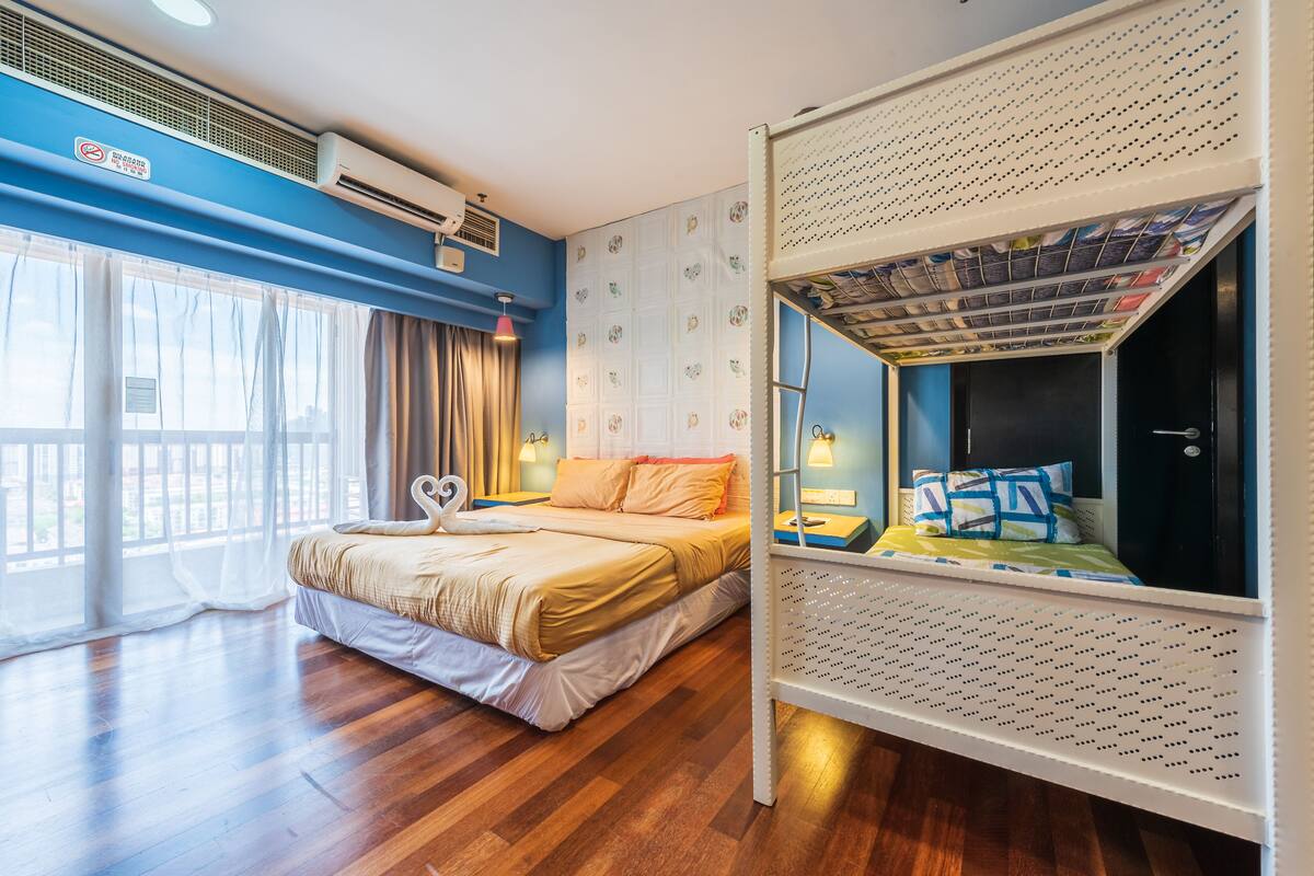A hotel room with a bed, a bunk bed, a window with curtains, an air conditioning unit, and a wall-mounted shelf with magazines.