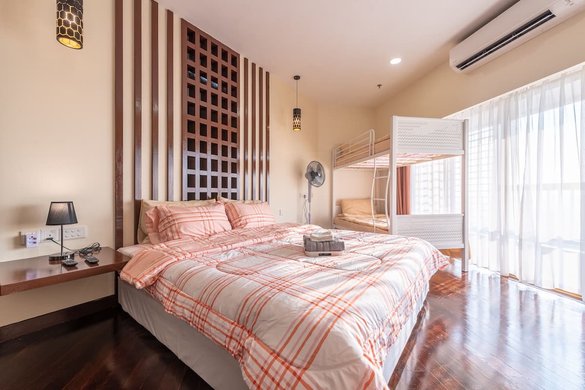 A bedroom with a large bed, a bedside table with a lamp, a fan, and a bunk bed in the background.