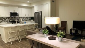 A modern kitchen with white cabinets, a microwave, and a refrigerator. There is a dining area with a table and chairs, and a TV in the background.