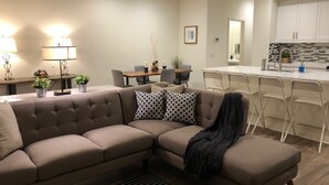 A modern living room with a sectional sofa, a dining area with a table and chairs, and a kitchen with white cabinets and a tiled backsplash.
